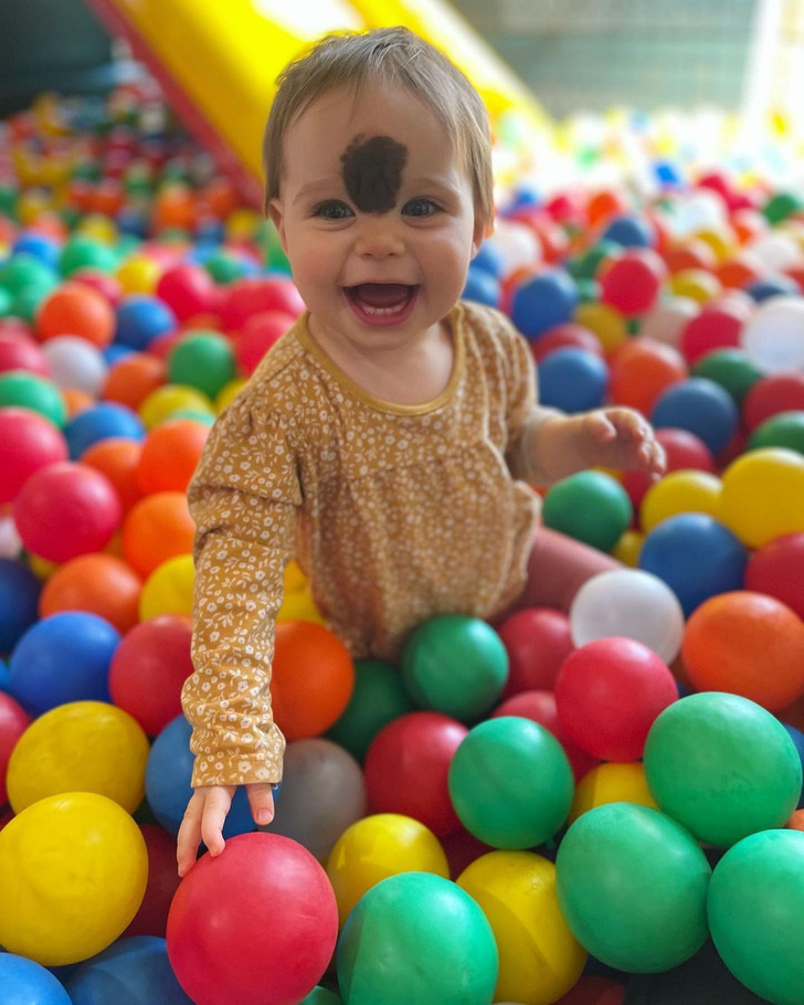 A baby girl with a brown birthmark on her forehead playing on a balls pool.